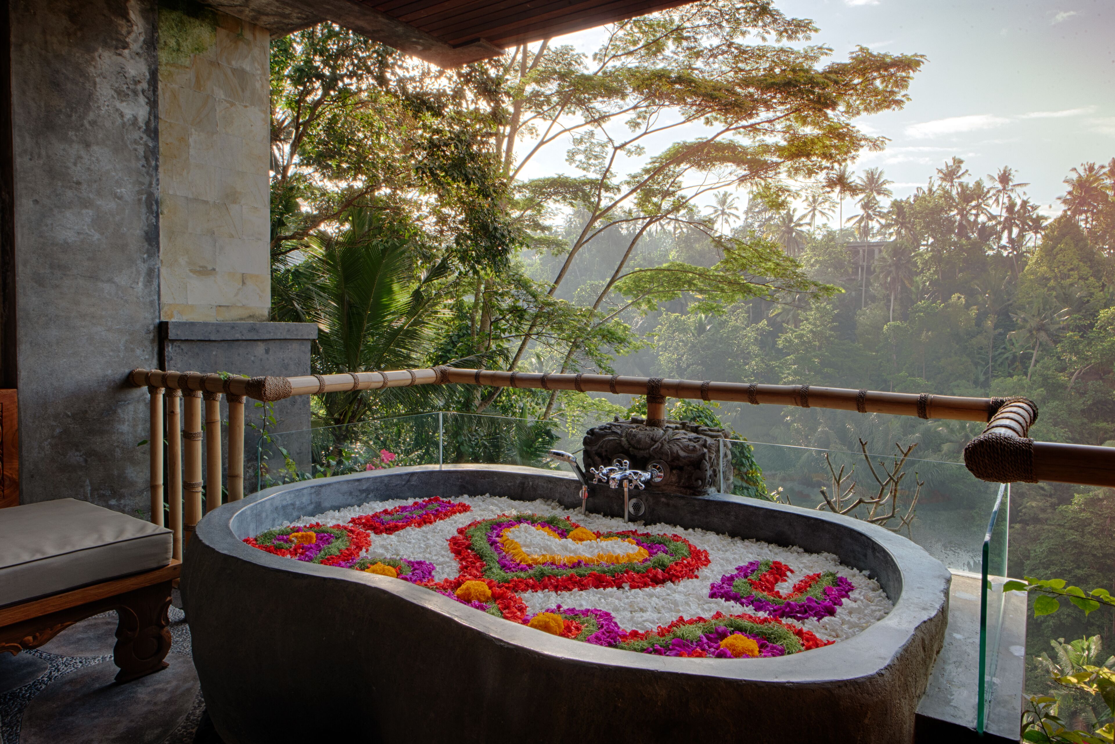 A stone bathtub filled with colorful flowers, a wooden bench, and a view of lush greenery and palm trees.