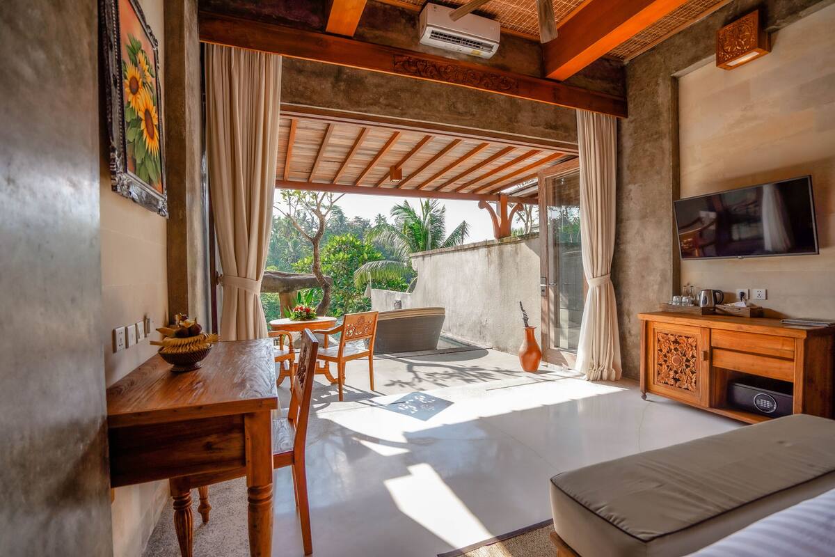 A room with a wooden dining table, chairs, a sofa, a TV, and a view of greenery outside.