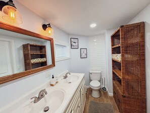 Baño con lavamanos doble, inodoro y espejo. Hay cestas de mimbre en la pared y un marco de fotos.