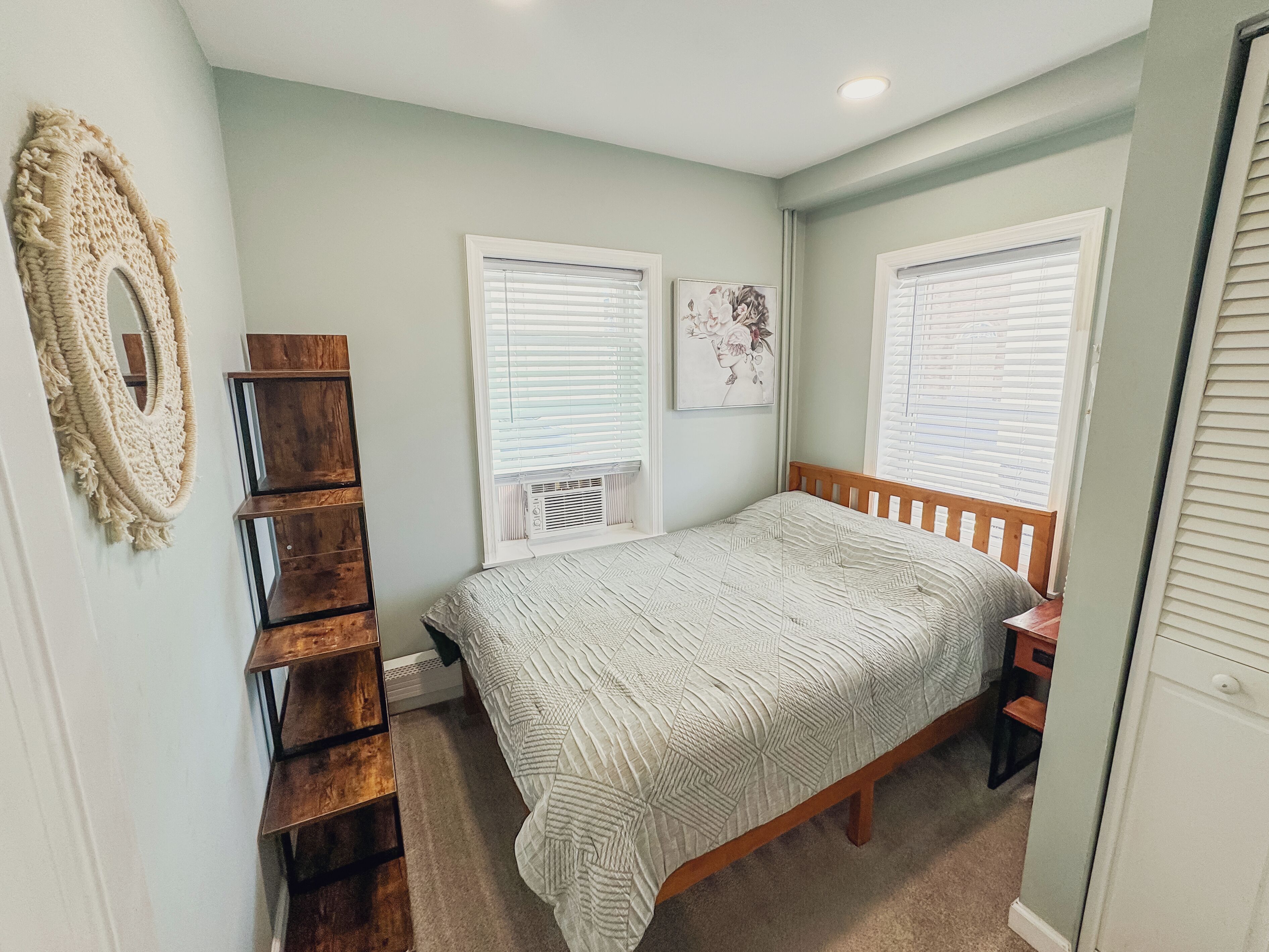 A bedroom with a bed, a wooden dresser, a macrame wall hanging, and two windows with blinds.