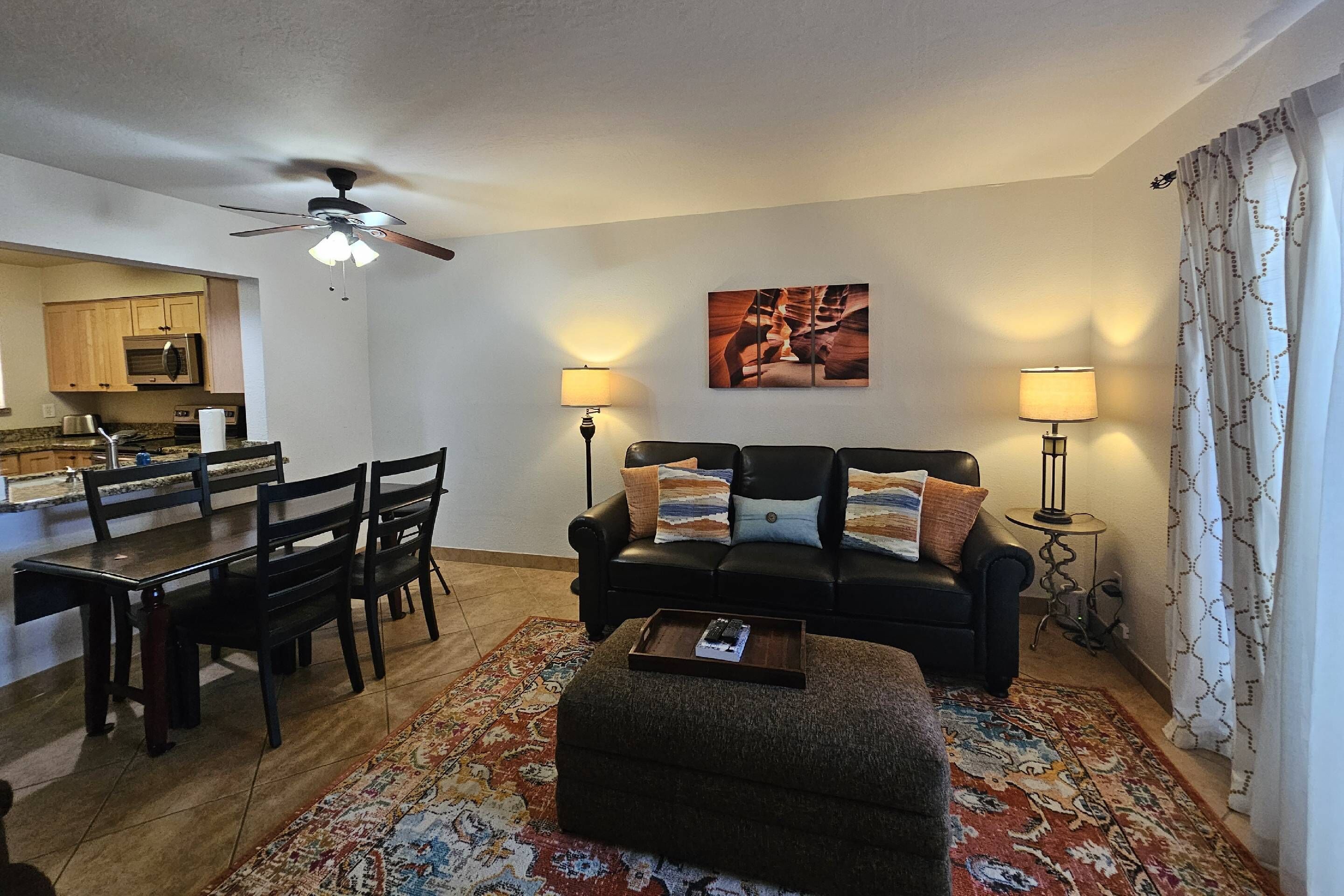 A living room with a black sofa, two lamps, a ceiling fan, a dining area with a table and chairs, and a kitchen in the background.