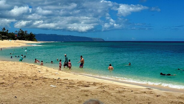 Una playa con agua turquesa cristalina, gente nadando y caminando por la orilla, y un cielo despejado con nubes dispersas.