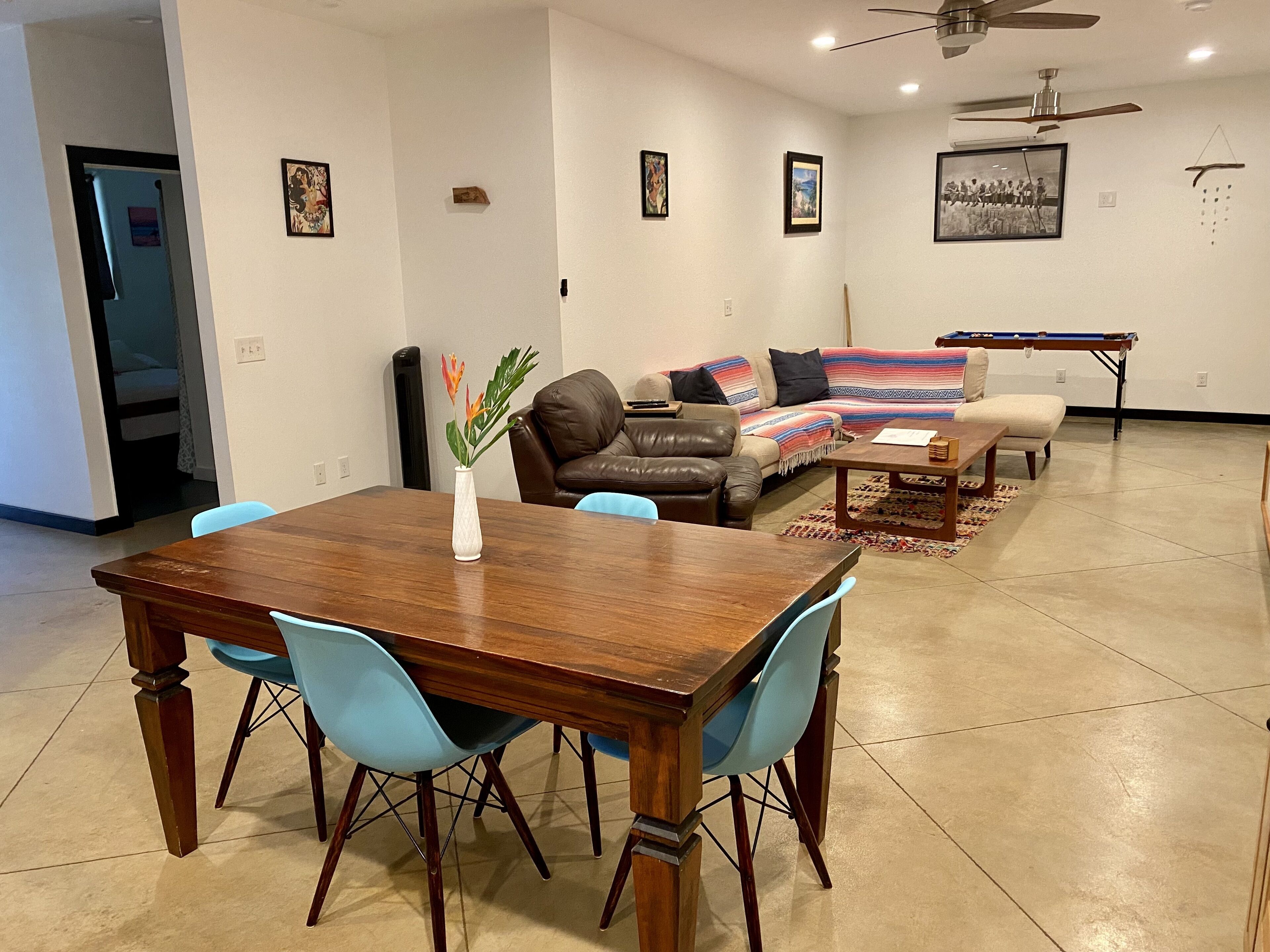A dining area with a wooden table and blue chairs, a sofa with a colorful blanket, and a ceiling fan.