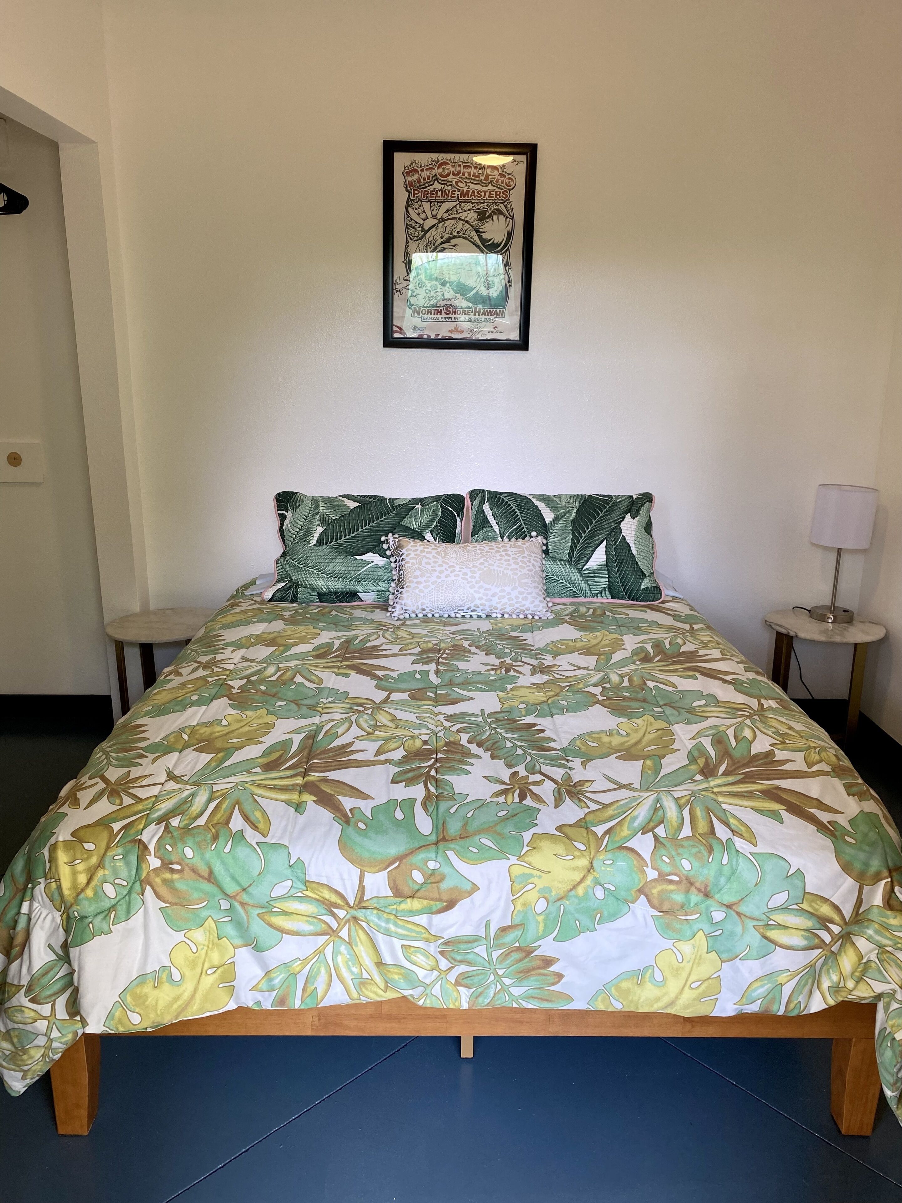 A bed with a tropical leaf pattern bedspread, a framed picture above it, and a small bedside table with a lamp.