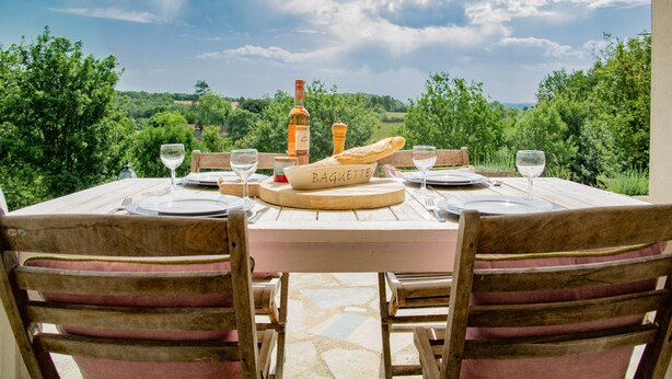 An outdoor dining table with a view of the countryside, a bottle of wine, and a baguette on the table. - Fine studio with large terrace and views (Cajarc)