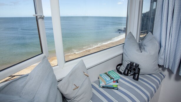 Une pièce avec une grande fenêtre donnant sur une plage. Sur le rebord de la fenêtre, on trouve des coussins et un coussin à rayures. Un livre et des jumelles sont posés sur le coussin.