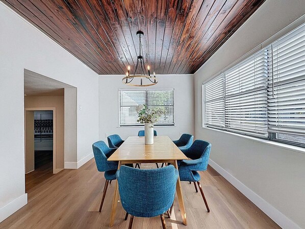 Un coin repas avec une table en bois, des chaises bleues, un lustre et une vue à travers les stores.