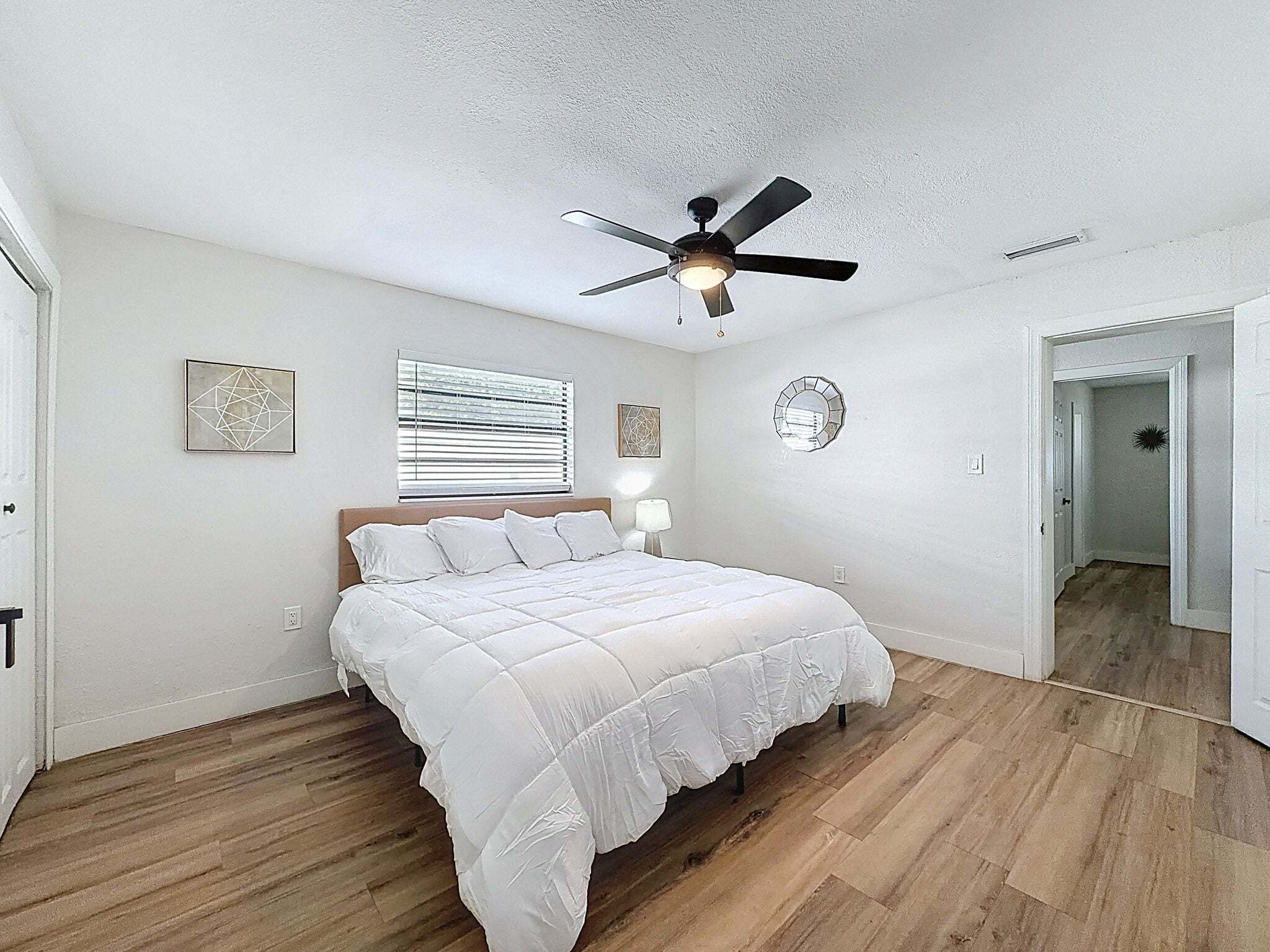 A bedroom with a bed, ceiling fan, artwork, window blinds, and a door leading to another room.