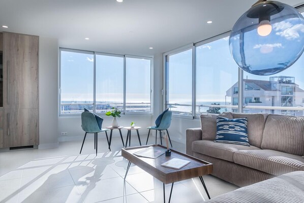 A modern living room with a large window, a sofa, a coffee table, and a dining area with chairs.