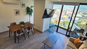 A modern living room with a dining area, large windows offering a city view, and a flat-screen TV mounted on the wall.