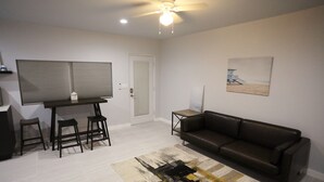A living room with a brown leather sofa, a coffee table, and a patterned rug. There is a door with a window and a painting on the wall.