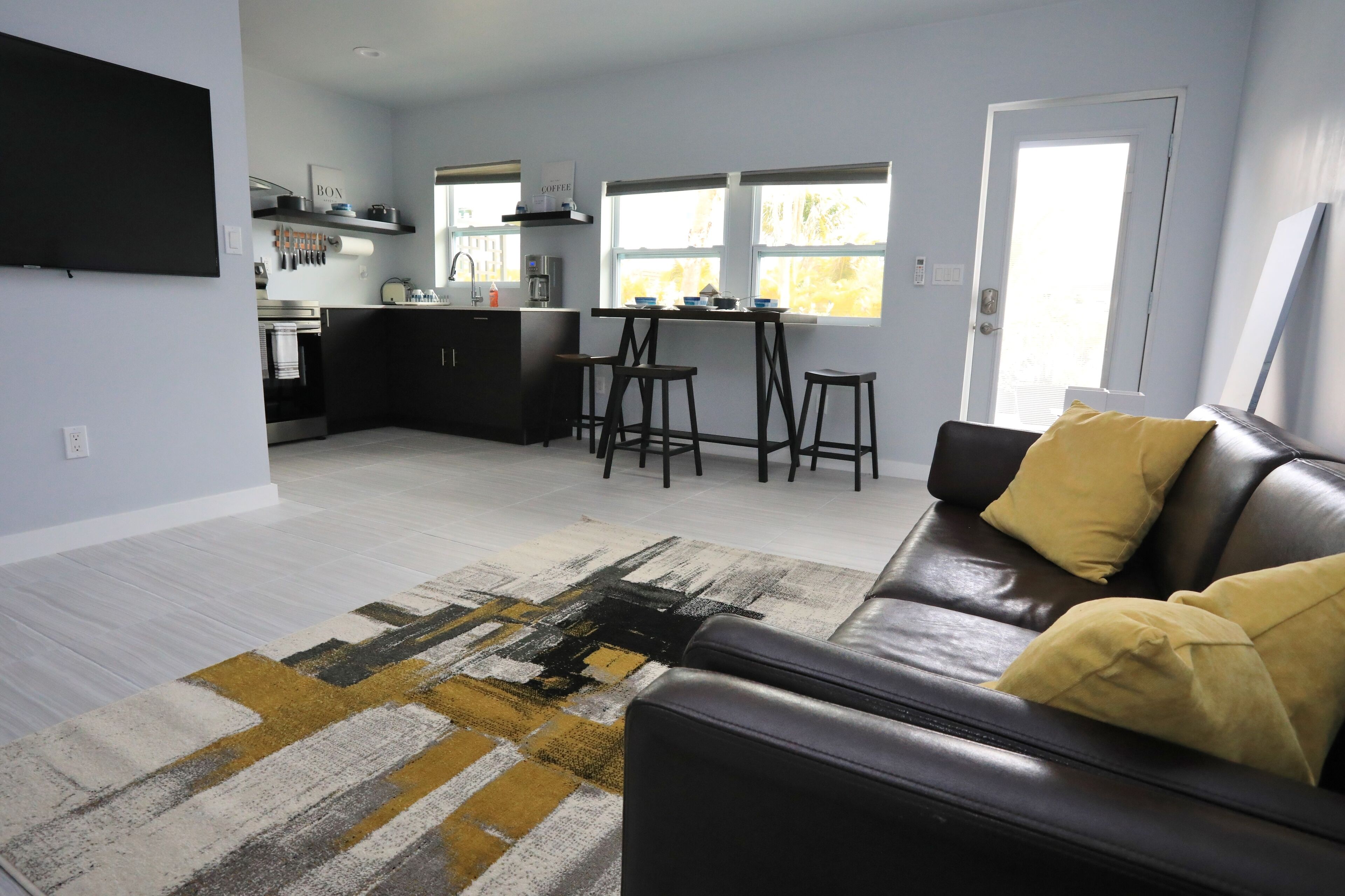 Un salon moderne avec un canapé en cuir noir, un tapis à motifs et une cuisine en arrière-plan.
