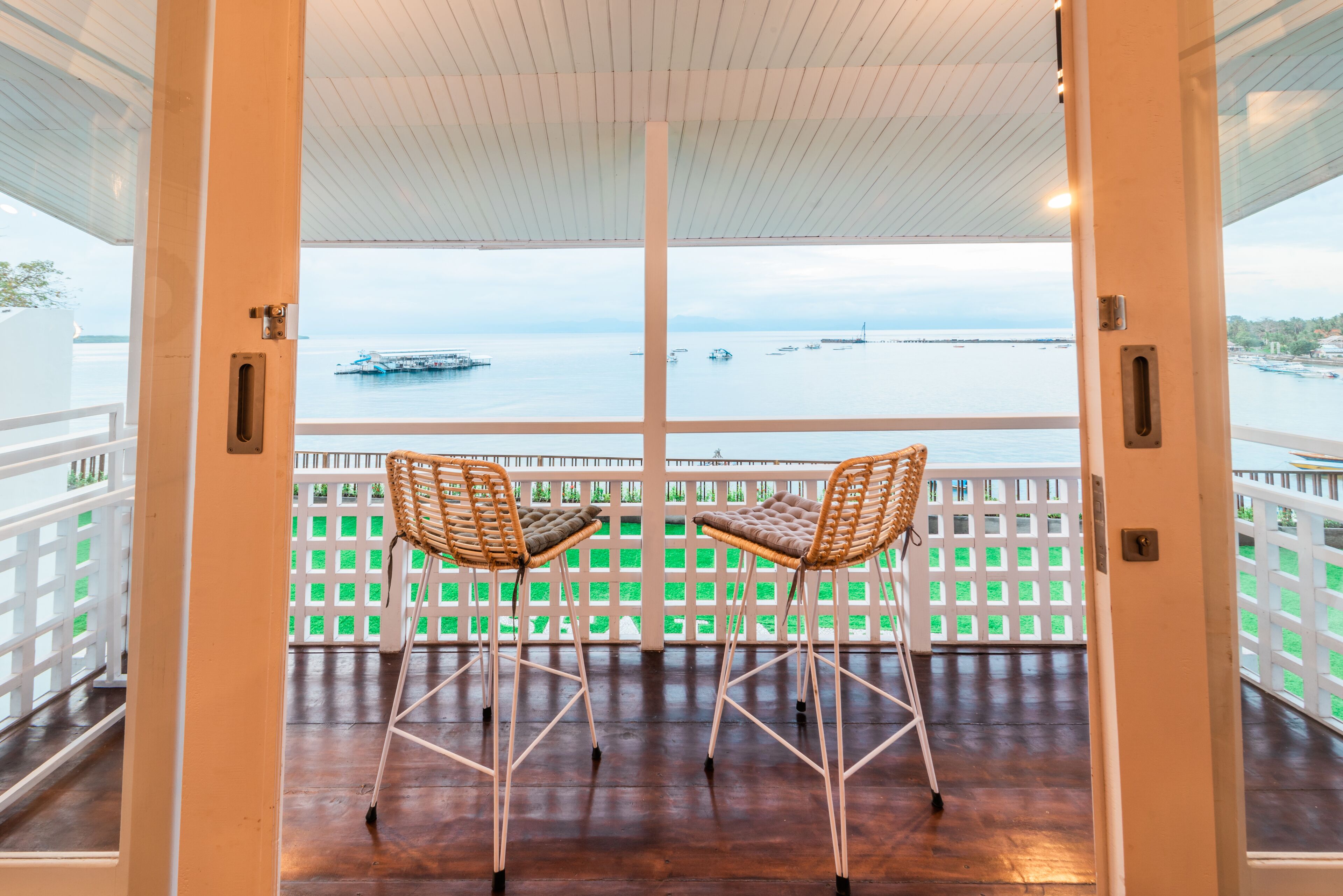 A balcony with two wicker chairs and a table, overlooking a calm sea with boats.