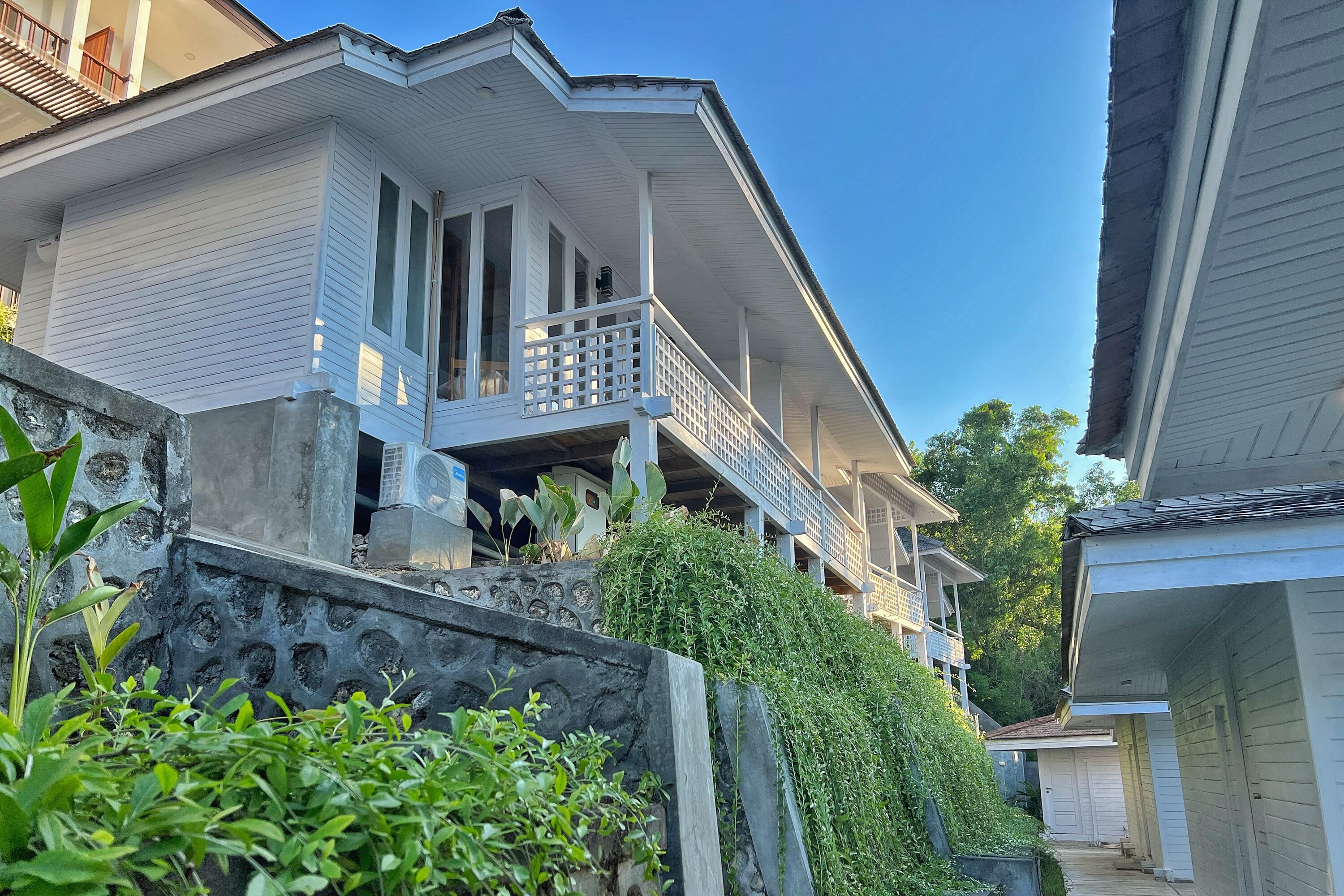 A two-story house with a balcony, a stone wall, and lush greenery.
