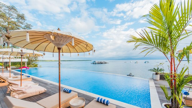Un espace aménagé au bord de la piscine, avec des chaises longues et des parasols, donnant sur une étendue d’eau calme où se trouvent des bateaux.