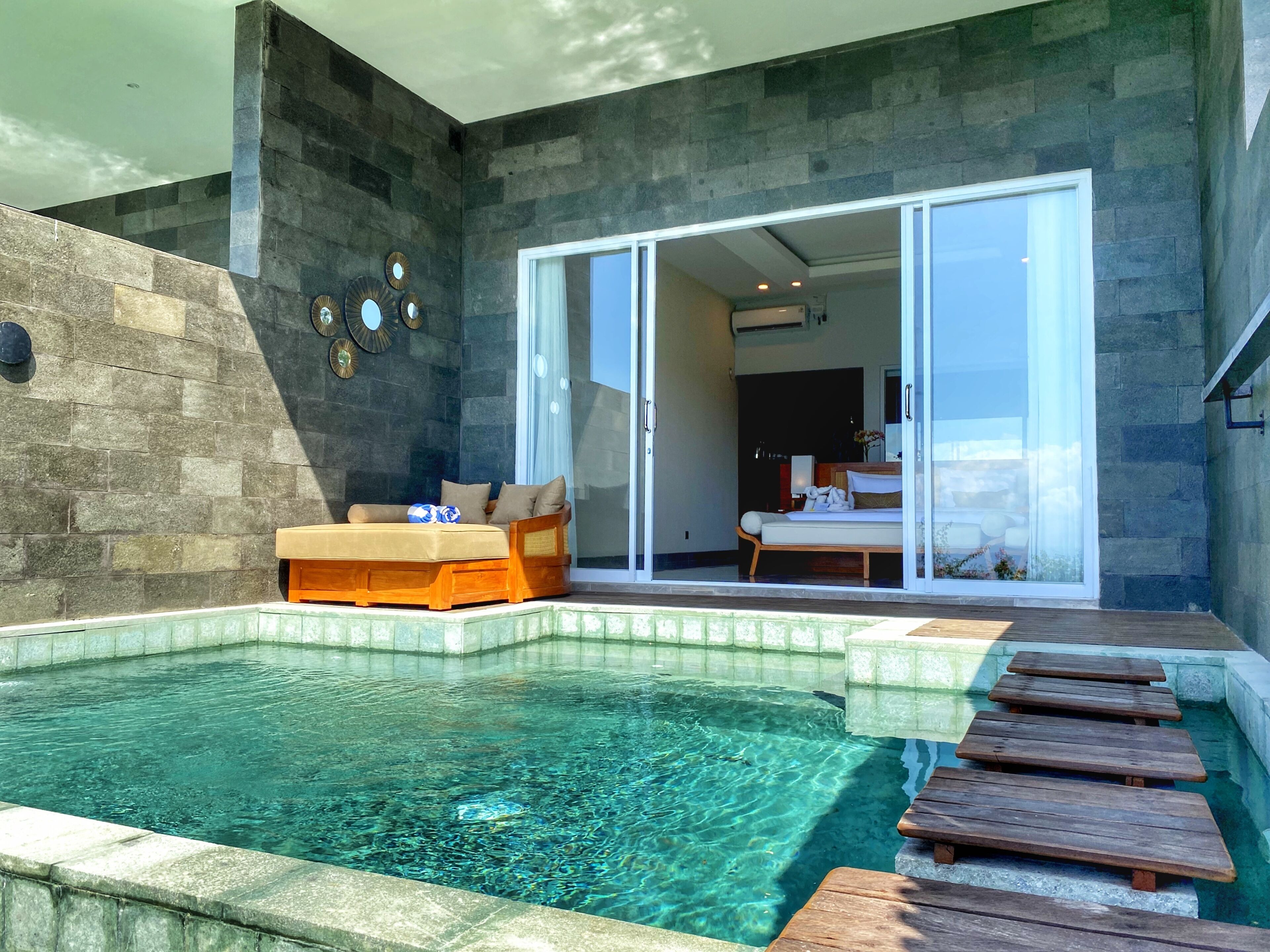 A modern outdoor pool area with a wooden deck, a lounge chair, and a glass door leading to a bedroom.
