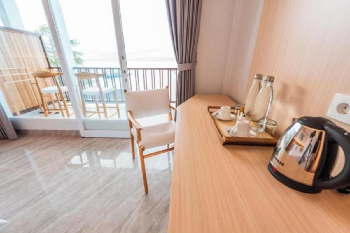 Une chambre d’hôtel avec un bureau en bois, une bouilloire et un plateau avec des tasses et un verre. Il y a une chaise et une vue sur l’extérieur, visible à travers une porte vitrée.
