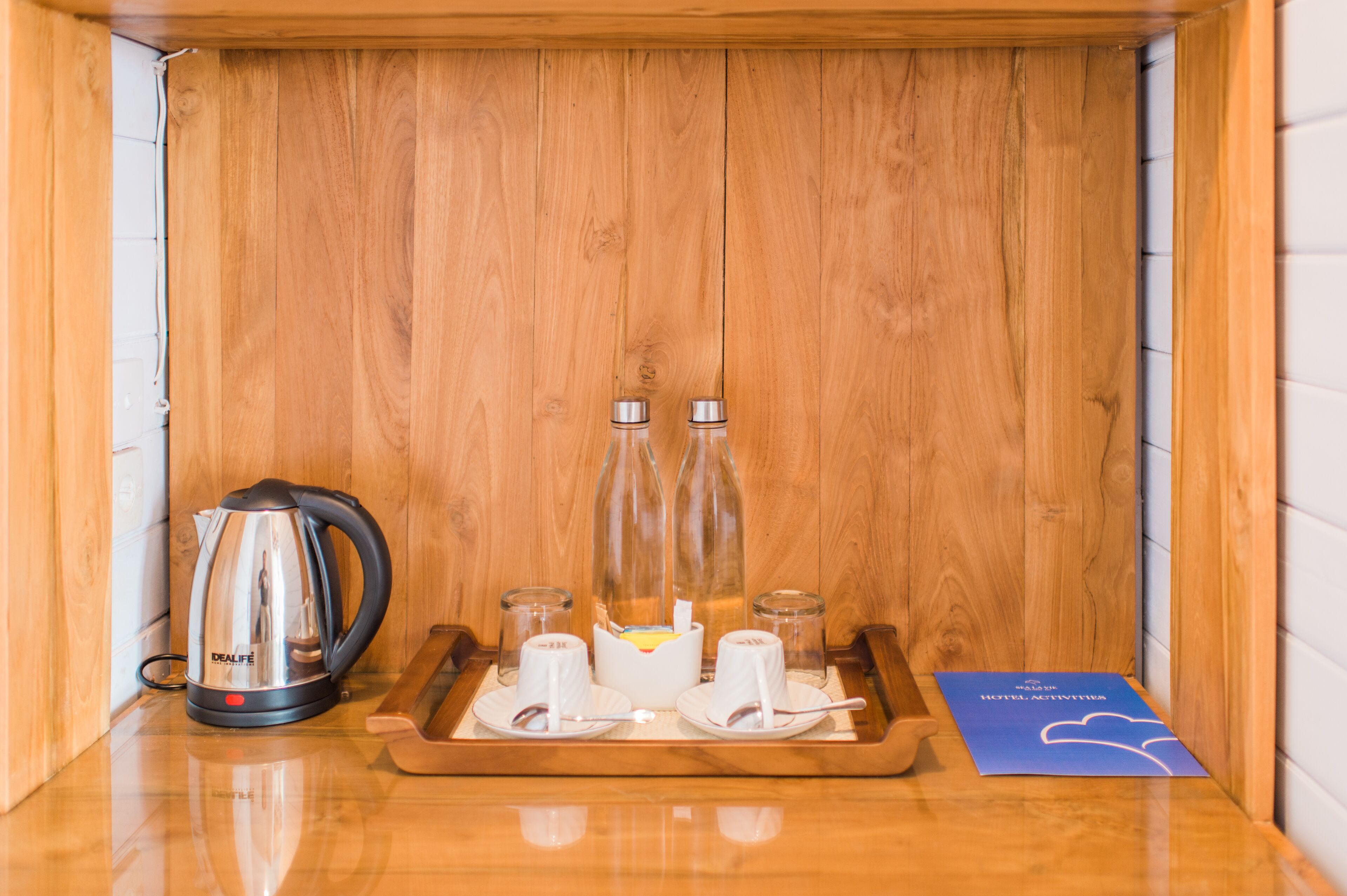 Un mueble de madera con un hervidor, vasos y una bandeja con tazas y un folleto.