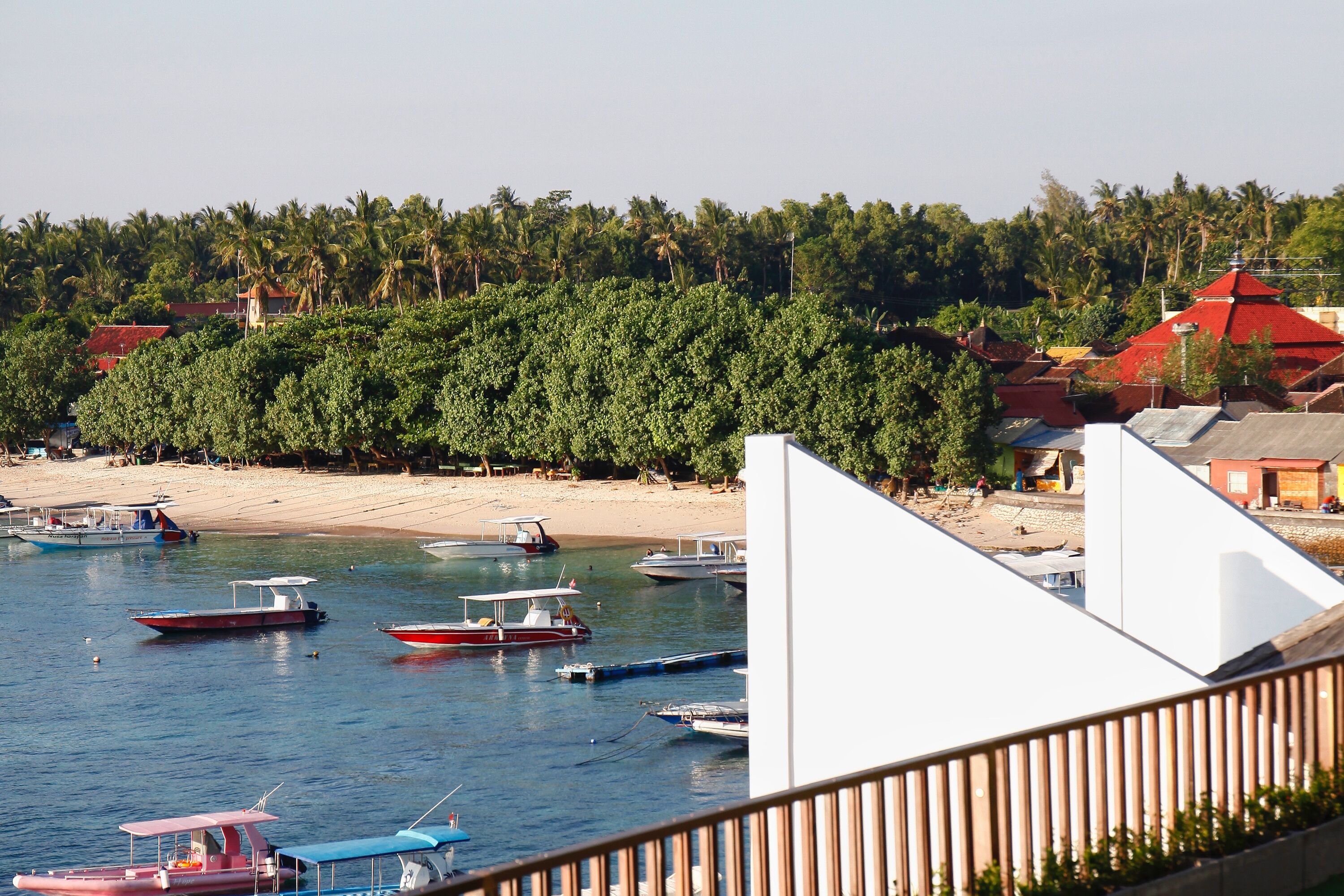 Una zona costera con barcos amarrados cerca de una playa arenosa, rodeada de abundante vegetación y edificios con tejados rojos.