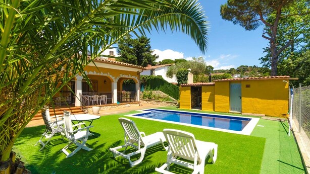 Zona de piscina con césped artificial, sillones blancos y un edificio amarillo con tejado de baldosas.