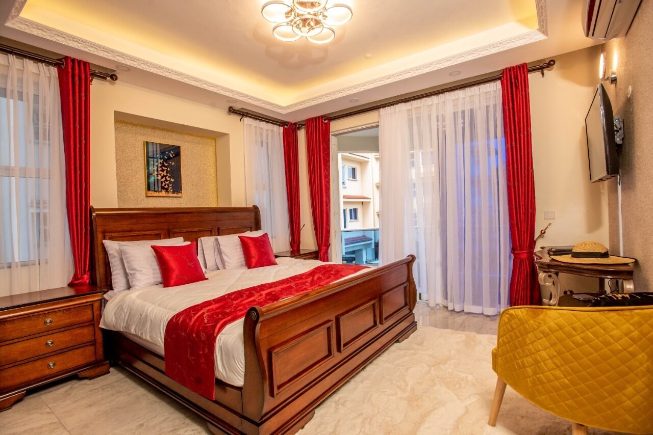 Een slaapkamer met een houten bed, rode gordijnen, een klein tafeltje en een gele stoel.