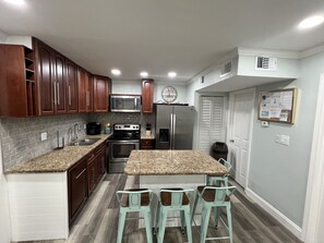 A compact kitchen with dark wooden cabinets, a granite countertop, a stainless steel refrigerator, and a microwave. There are three bar stools and a clock on the wall.