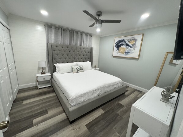 A bedroom with a large bed, a nightstand, a ceiling fan, and a painting on the wall.