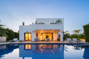 Una casa moderna de dos pisos con piscina delantera, que cuenta con un patio con sillas de descanso y un interior bien iluminado.