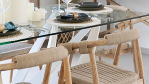 Een eettafel met glazen blad, omringd door rieten stoelen, gedekt met borden, glazen en kaarsen.