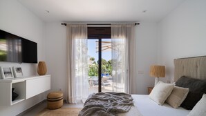 Een moderne slaapkamer met een bed, een aan de muur bevestigde televisie, een raam met dunne gordijnen en uitzicht op het omliggende landschap.