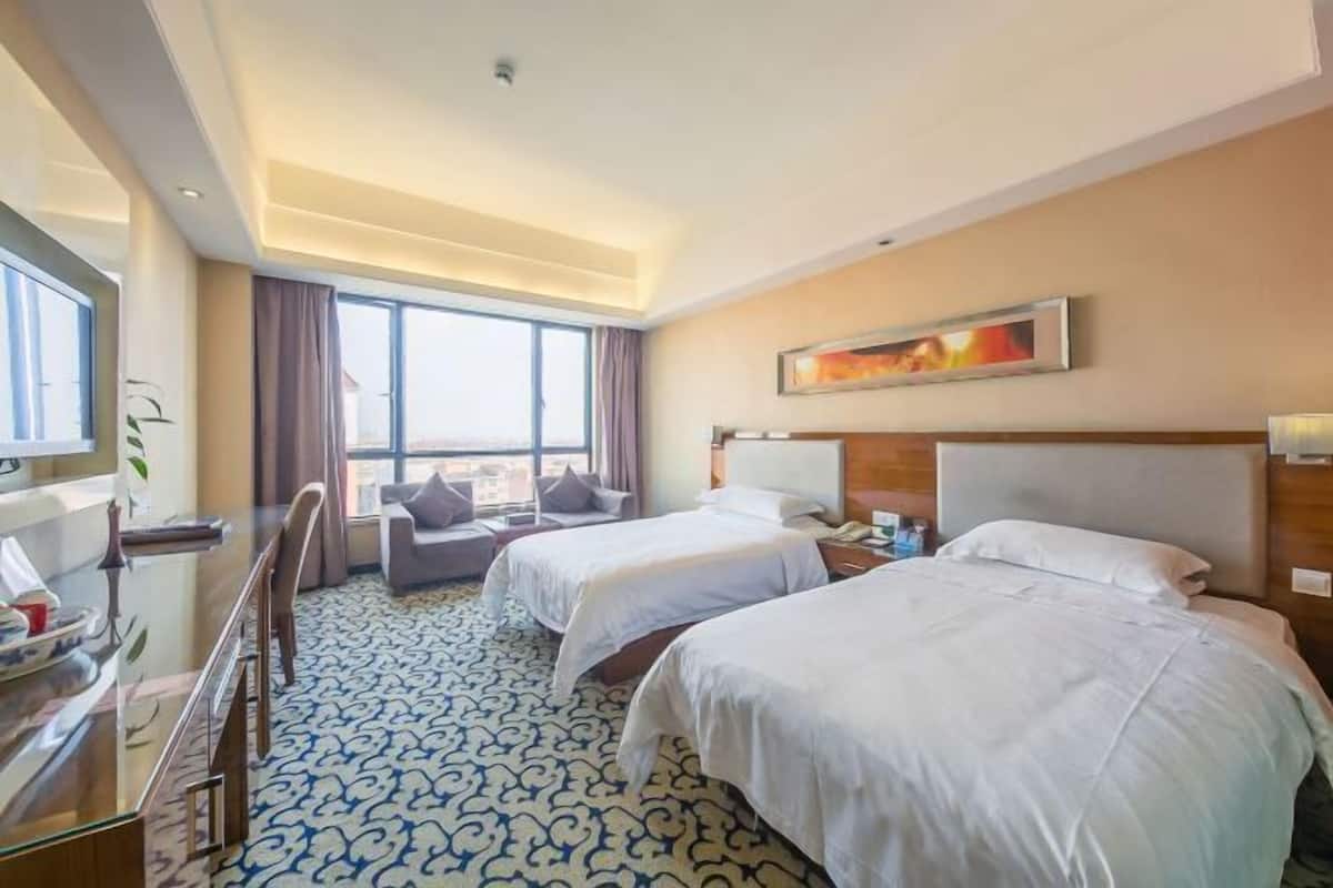 A hotel room with two beds, a desk, a chair, a TV, and a large window.