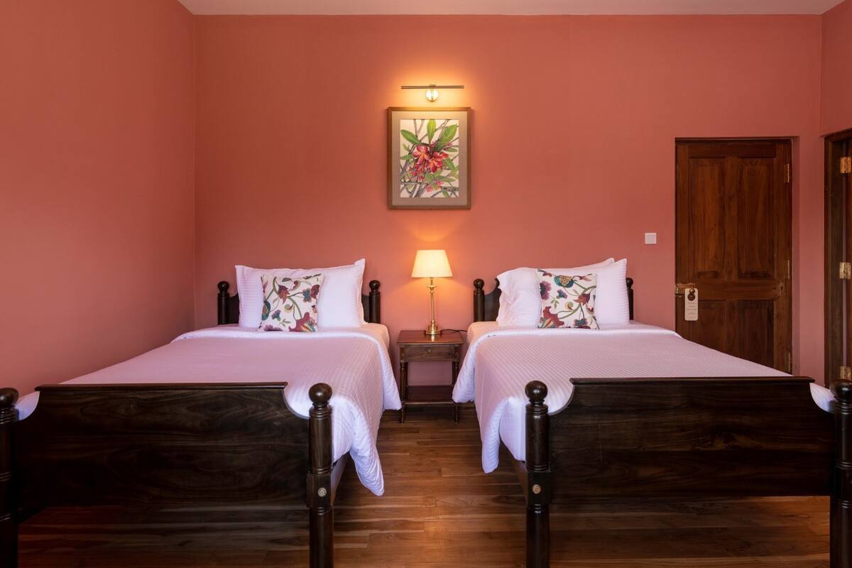 Una habitación con dos camas de madera, un mesita de noche con lámpara, una obra de arte floral enmarcada en la pared y una puerta de madera.