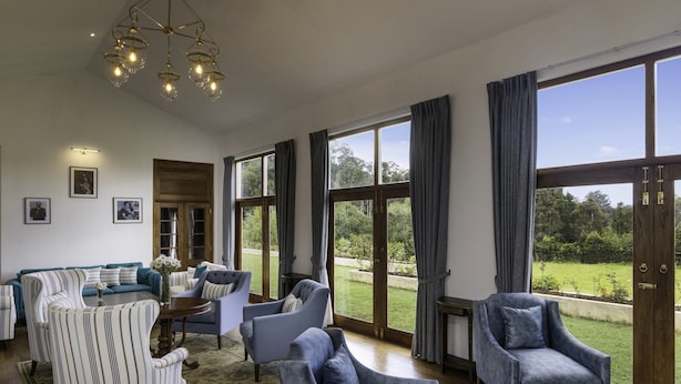 A spacious living room with a large sofa, armchairs, and a coffee table. There are large windows with curtains, a chandelier, and framed pictures on the wall.