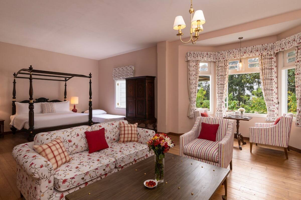 Un dormitorio con una cama con dosel, un sofá, un sillón a rayas, una mesa de centro de madera con un florero y vistas a la vegetación a través de la ventana.