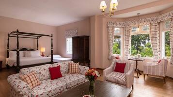 Un dormitorio con una cama con dosel, un sofá, un sillón a rayas, una mesa de centro de madera con un florero y vistas a la vegetación a través de la ventana.