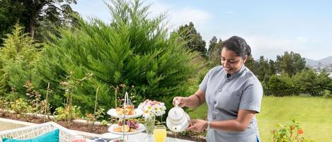 Una persona sirve té en una taza en una mesa al aire libre, con una bandeja de postres de tres niveles, rodeada de follaje y un cielo despejado.