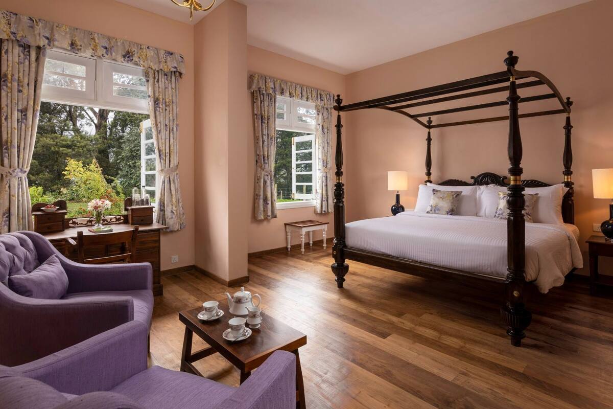 Una cama con dosel de madera, una mesita de noche con lámpara y una silla con una mesa dispuesta para té.
