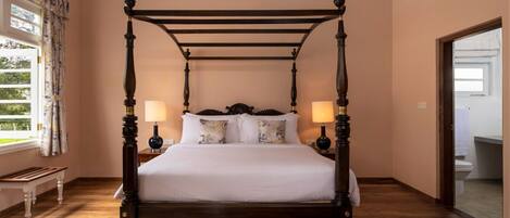 Una cama con dosel, ropa de cama blanca, dos mesitas de noche con lámparas, un banco de madera en una habitación con suelo de madera y una ventana con cortinas.