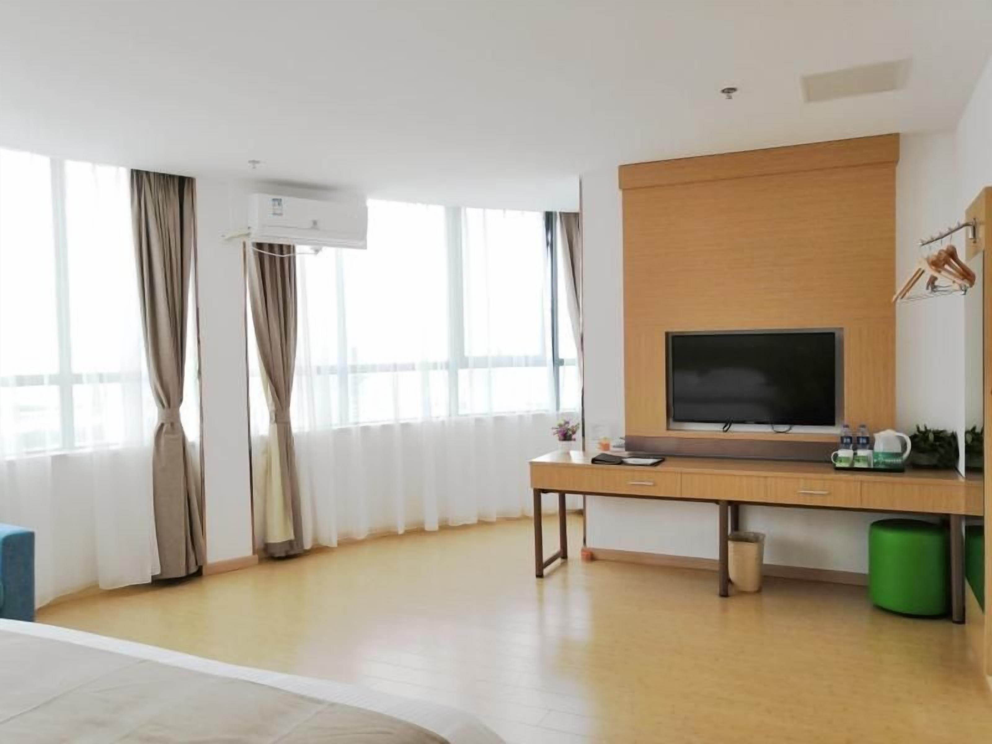 Quarto de hotel moderno com uma janela ampla, televisão de tela plana, escrivaninha de madeira e uma lixeira verde.