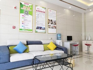 Ein Empfangsbereich mit einer Couch, einem Glastisch, einem Wasserspender und Informationsplakaten an der Wand.