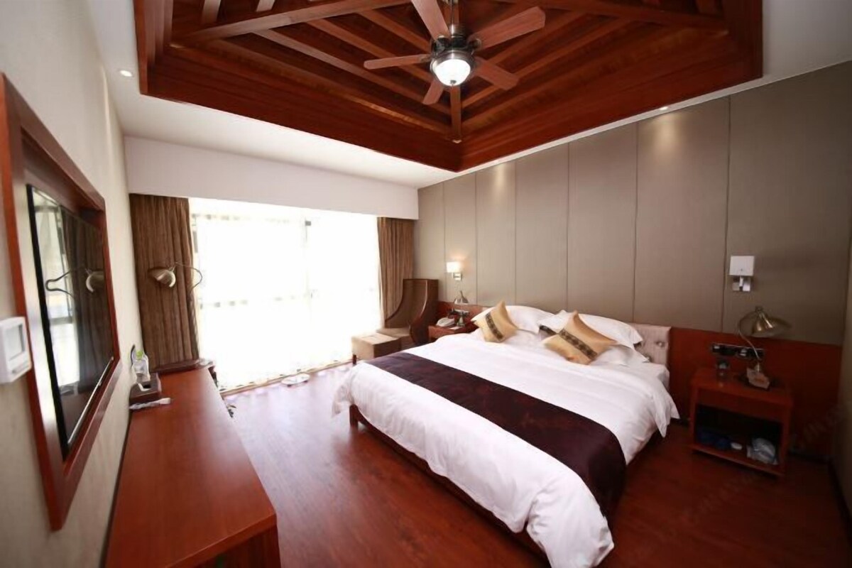 Una habitación de hotel con techo de madera, una cama grande con ropa de cama blanca y oscura, una mesita de noche con lámpara y una ventana con cortinas.