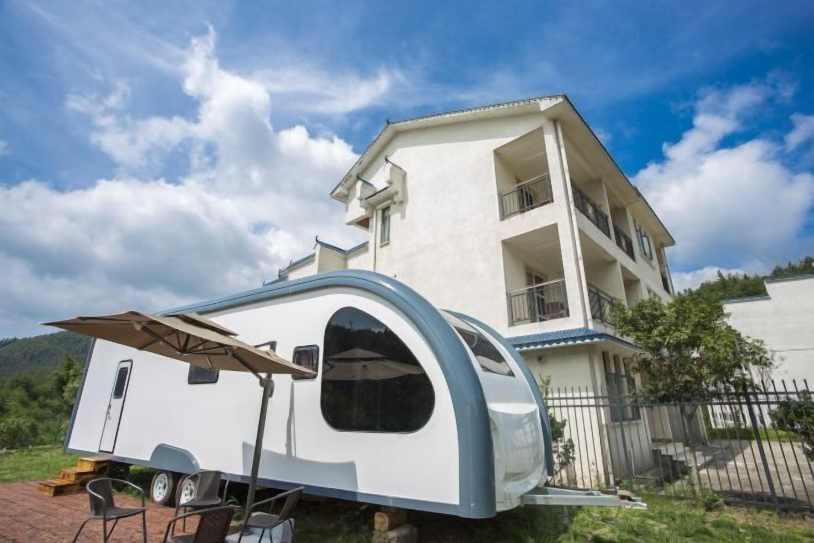A white two-story house with a balcony, a small attached trailer, and a clear blue sky with clouds.