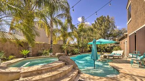 A swimming pool with steps leading into it, surrounded by palm trees and a patio area with lounge chairs and a table. - Family Gem in Phoenix w/ Saltwater Pool & Theater (Gilbert)