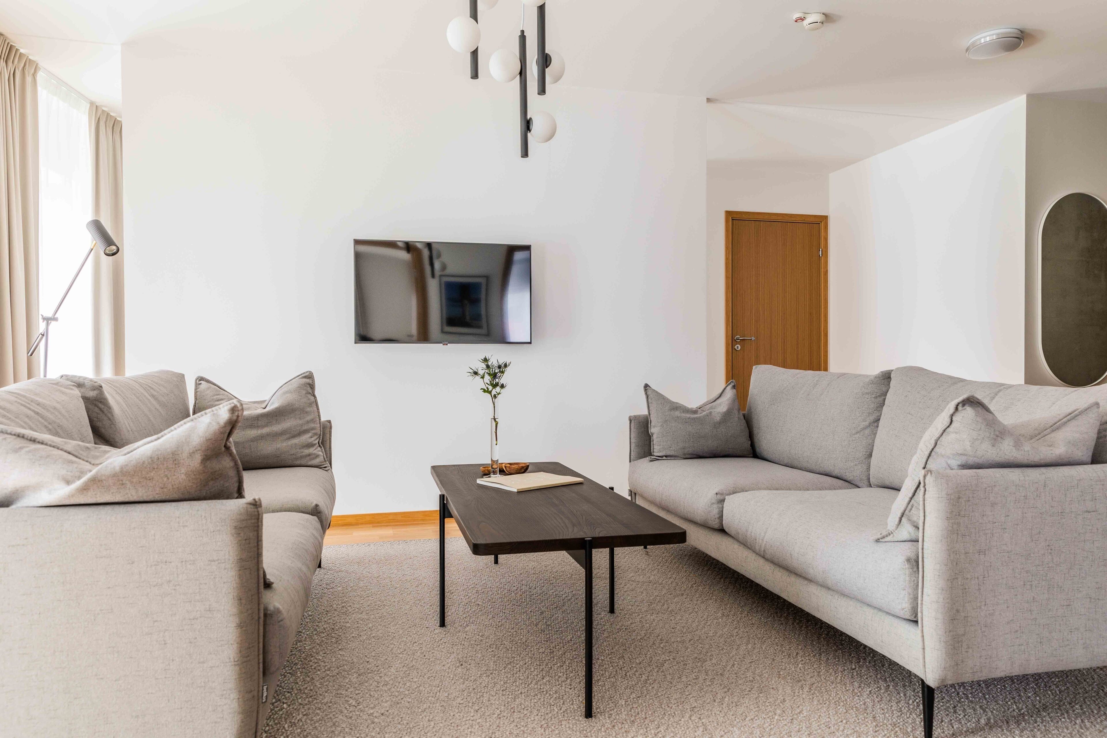 A modern living room with two grey sofas, a dark wooden coffee table, and a flat-screen TV mounted on the wall.