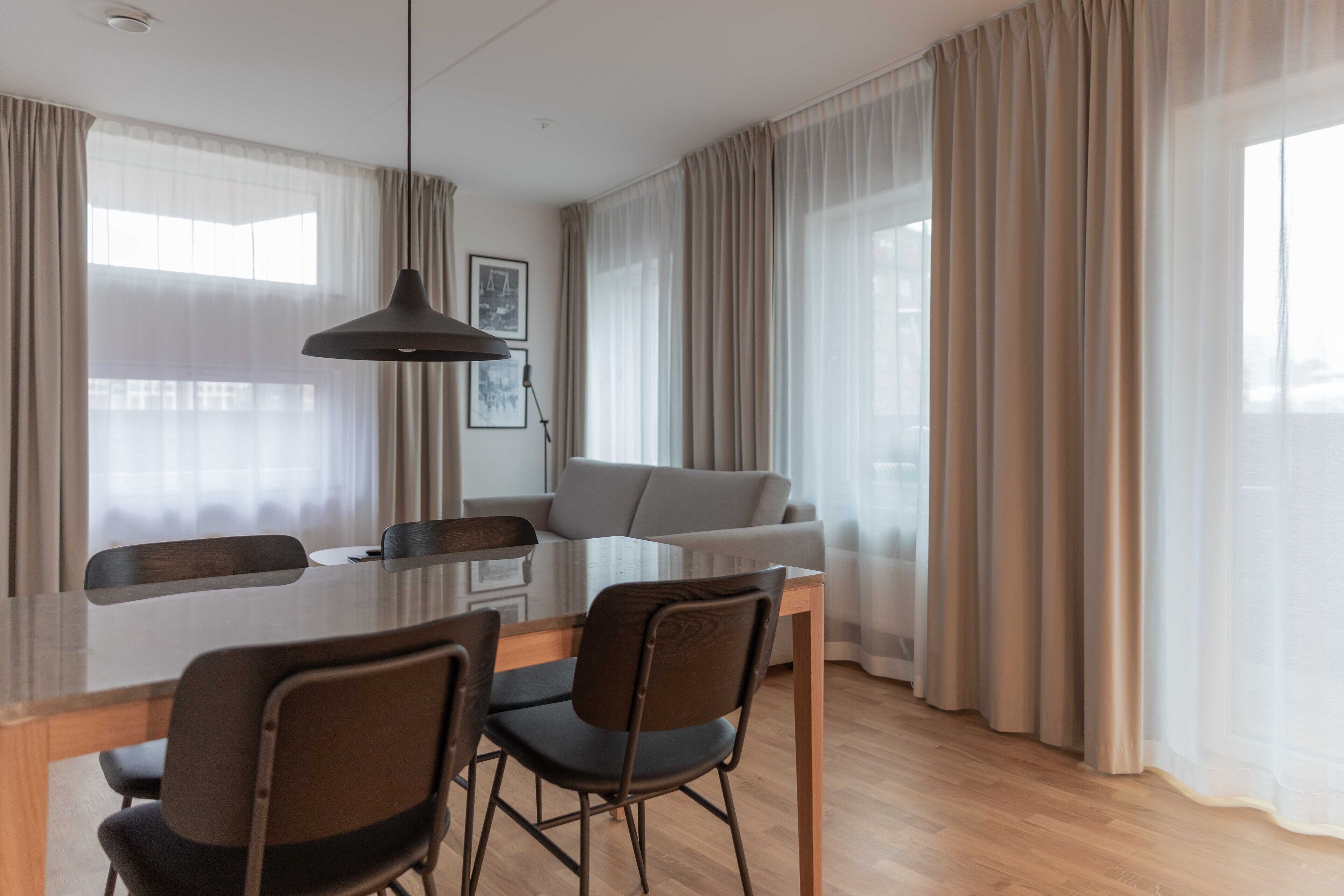 Una sala de estar moderna con comedor, que incluye una mesa de madera, dos sillas, un sofá y un ventanal con cortinas traslúcidas.
