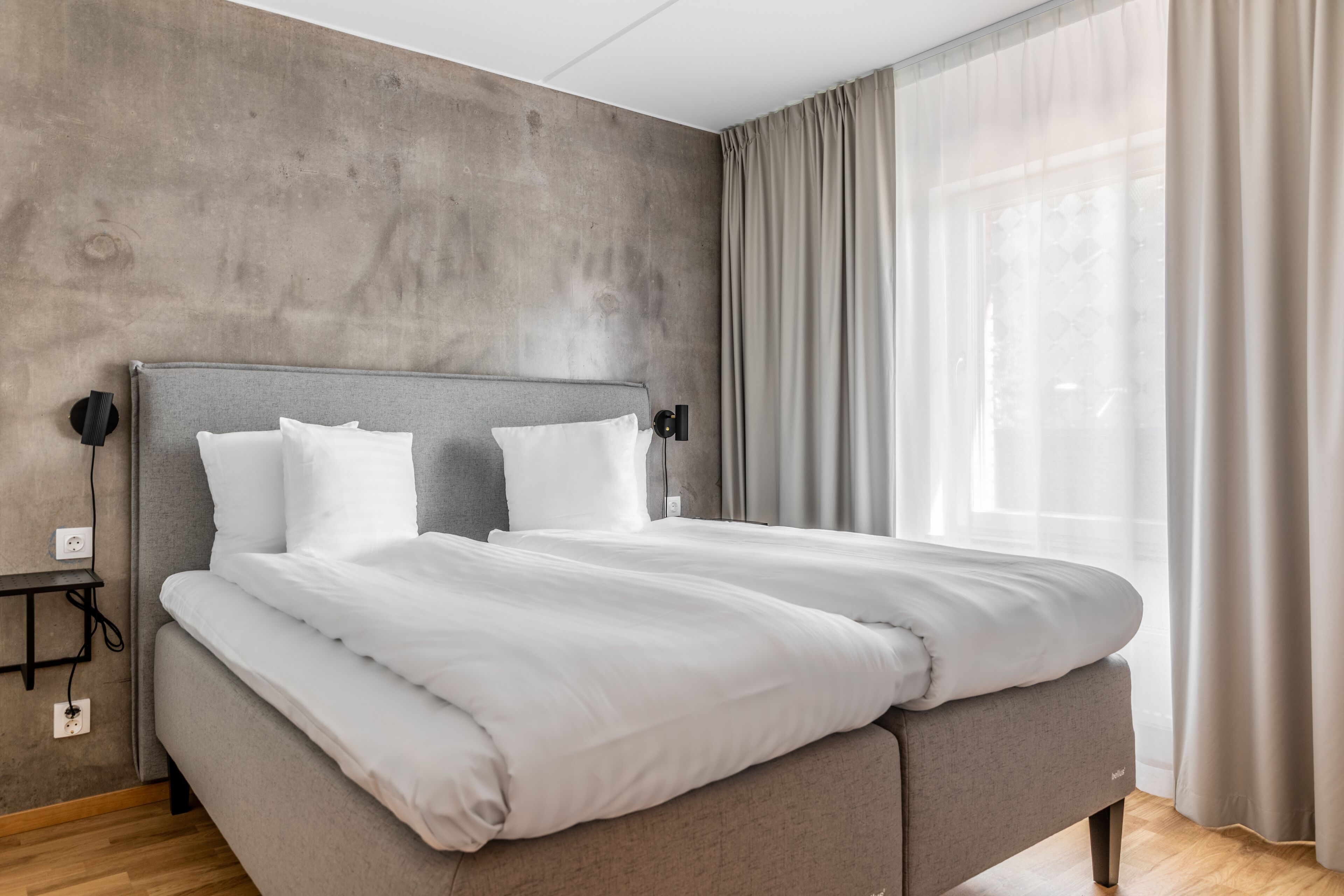 A modern bedroom with a grey headboard, white bedding, and a large window with sheer curtains.
