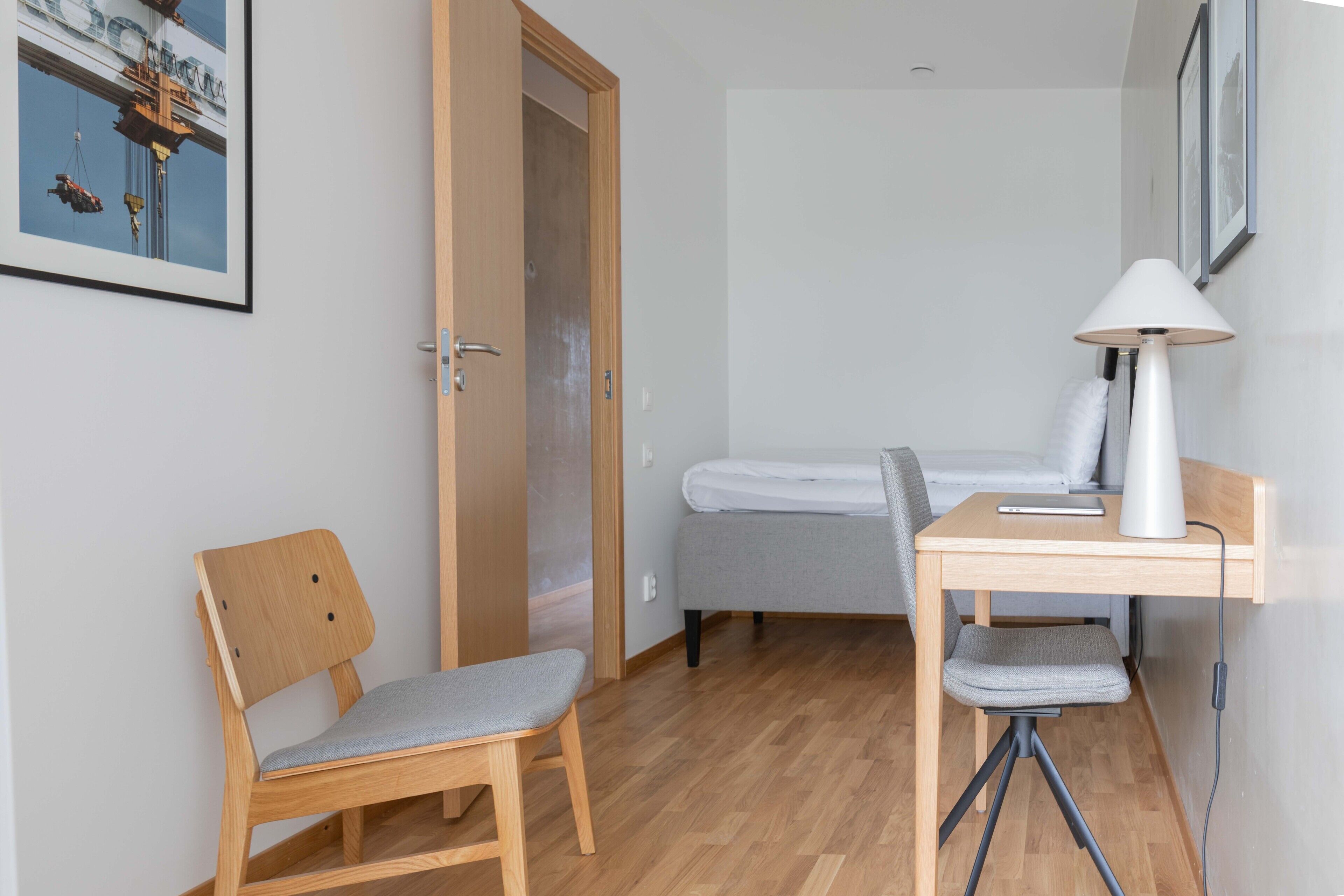 Een kleine, netjes ingerichte kamer met een houten bureau, een stoel, een bed en een lamp.