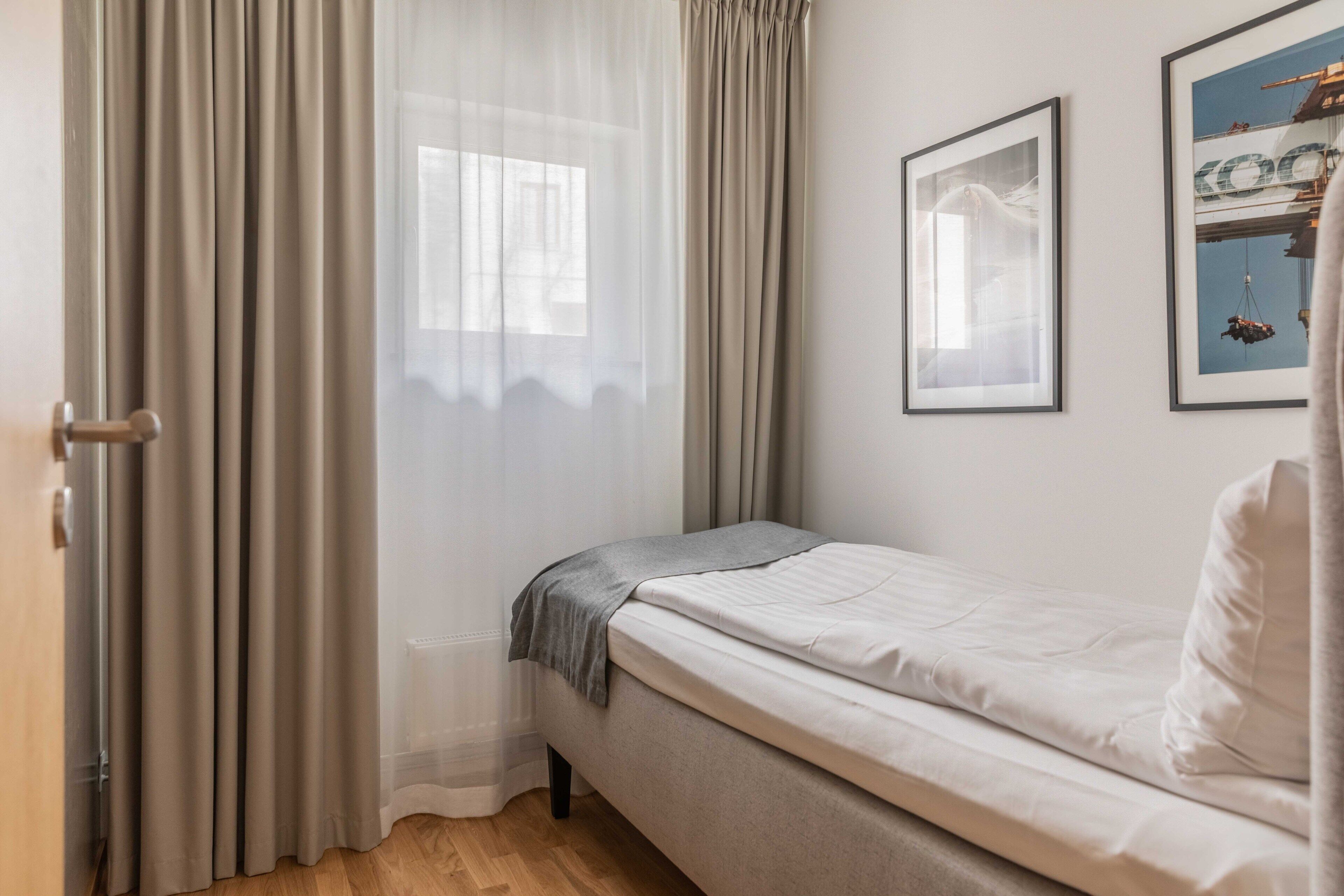 A bedroom with a bed, a window with sheer curtains, and two framed pictures on the wall.