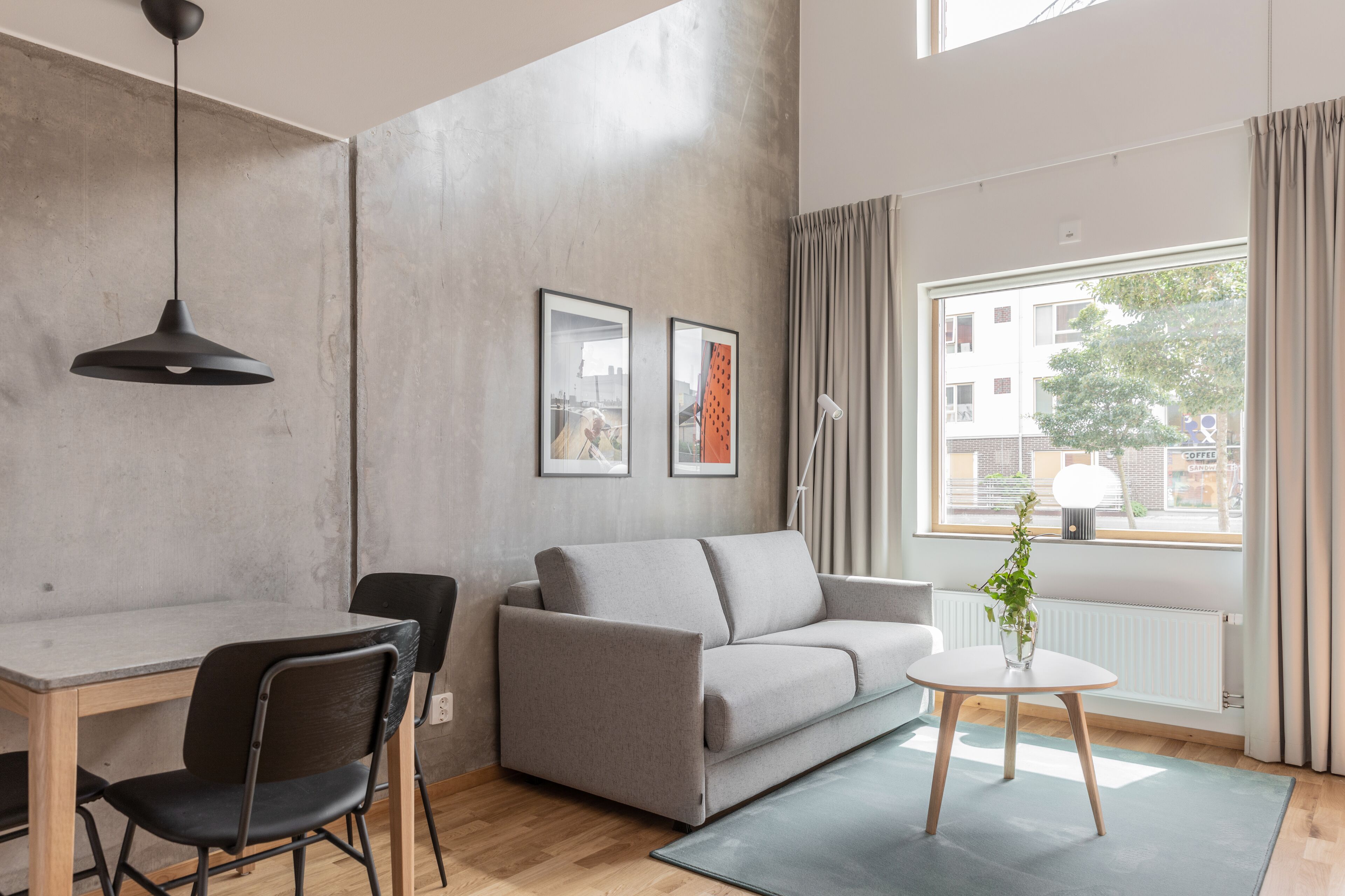 A modern living room with a grey sofa, a small round table, and a black chair. There are two framed pictures on the wall and a window with curtains.