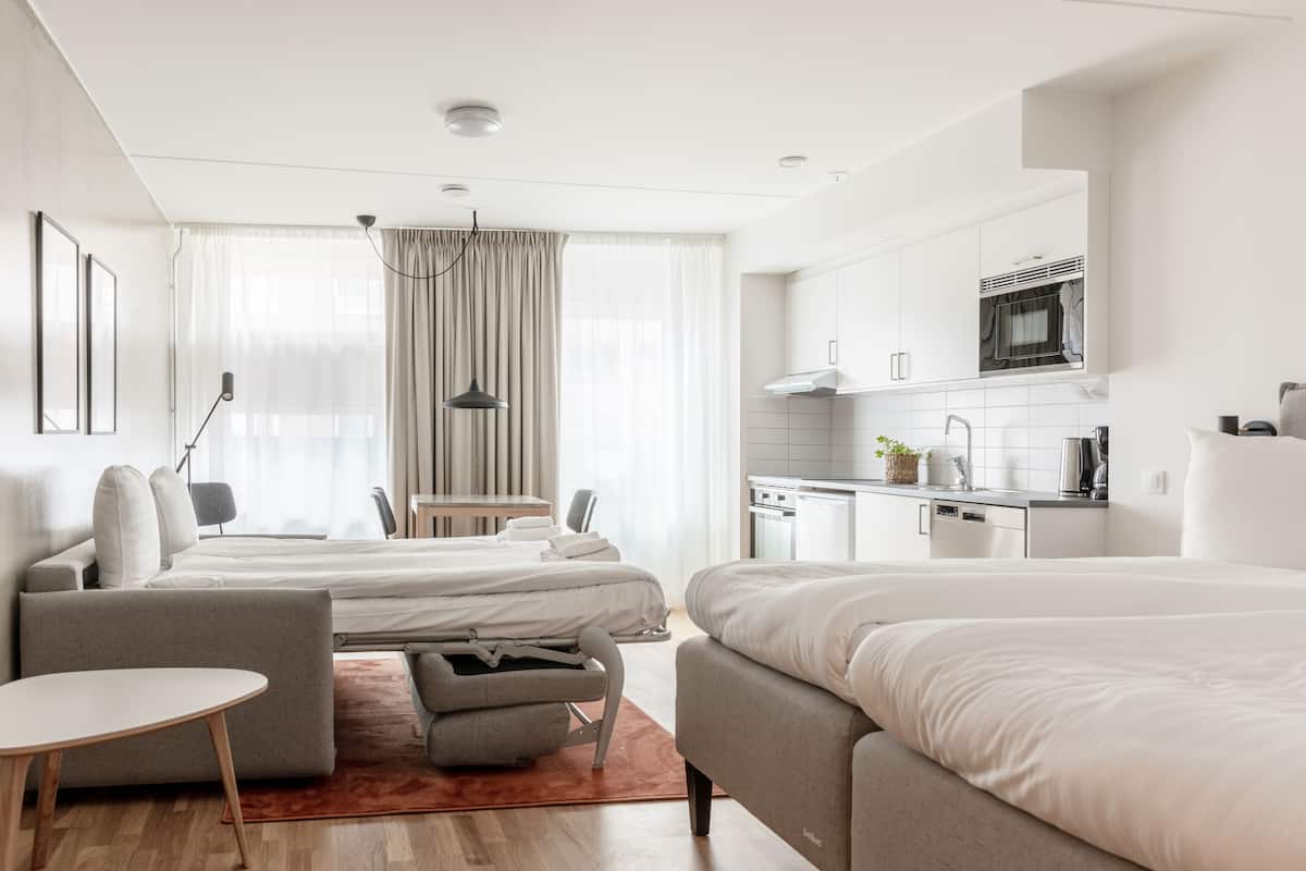Een moderne hotelkamer met twee bedden, een klein zitehoekje, een kitchenette en een groot raam met gordijnen.